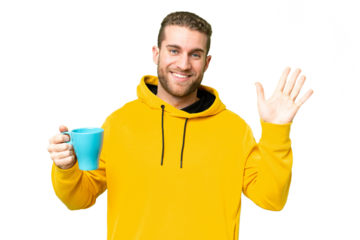 Young handsome blonde man holding cup of coffee over isolated background saluting with hand with happy expression