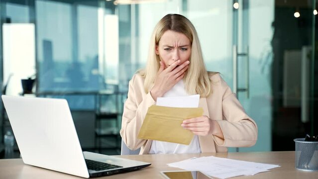 Sad depressed young businesswoman reading letter with bad news while sitting at workplace in modern office. Frustrated shocked blonde female employee in a suit looking at unpleasant notification