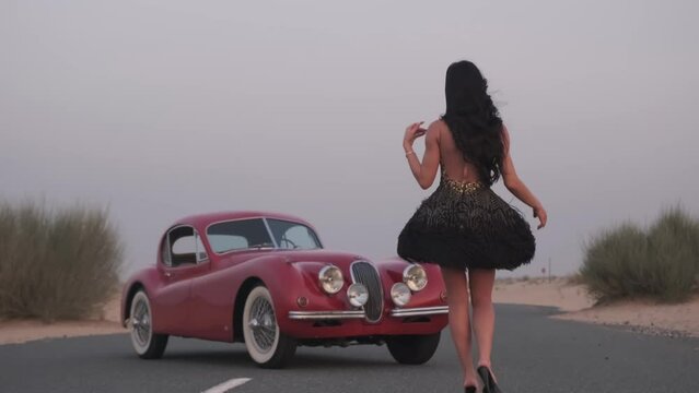 Young woman in a black dress, with long hair and bright makeup. He walks in high-heeled shoes on the road to a red retro car.