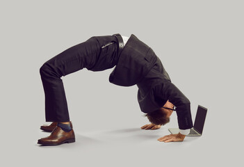 Funny energetic businessman in suit practises yoga while working on his modern laptop computer. Fit...