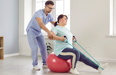 Male physiotherapist helps female patient do exercise for leg muscles. Fat woman sitting on Swiss fitball and doing sports exercise with elastic band. Physiotherapy concept