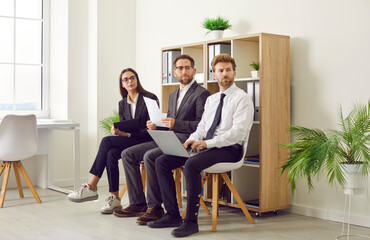 Young business people sitting on the chairs in a row with resumes and laptops in hands. Group of a staff and company employees. Job candidates seekers waiting for interview invitation turn.