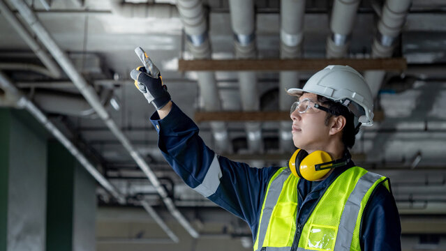Portrait of confident man construction worker with reflective vest, safety helmet, protective goggles and ear muffs. Asian male engineer pointing at piping system at construction site