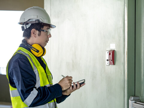 Building inspector man using digital tablet checking wall mounted fire alarm station near fire exit door. Asian male worker in reflective vest and helmet checking door hardware for safety and security