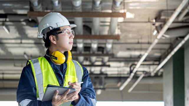 Building inspector man using digital tablet checking plumbing pipeline system. Asian male worker in reflective vest, ear muffs, safety helmet and goggles working for building maintenance inspection