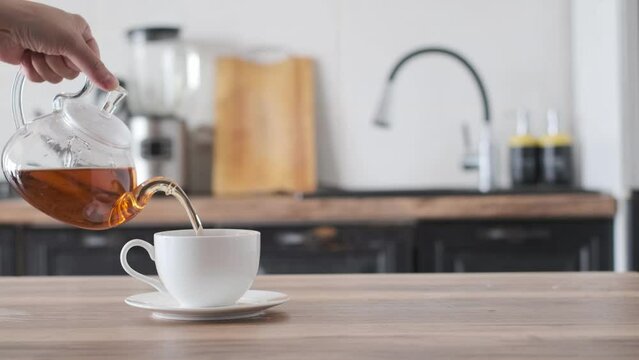 Hand pouring tea from glass tea pot to the cup on a home kitchen background