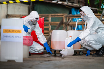 A team of two chemists, wearing PPE suits and gas masks, recover a deadly chemical spill on the factory warehouse floor. Correct disposal of chemical spills in industrial plants.