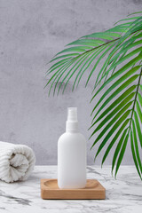 White cosmetic bottle with spray pump with towels and a palm leaves on a wooden table and concrete wall. Sun protection and sunscreen blank product. Beauty skin care product template.