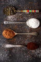 Spoons with green tea, cocoa, icing sugar and ground coffee on black background sprinkled with...