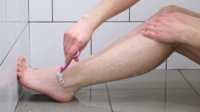A woman shaving her legs with a pink razor sitting in the shower. Hairy shins. hygiene procedures. Body care. Smooth white skin. Close-up. Hygiene and cosmetics concept.