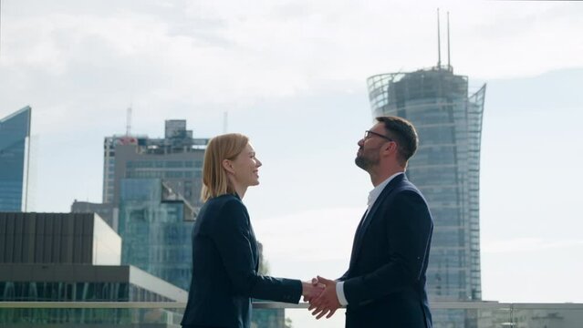 Side view two successful business partners male female handshaking meet and shake hands coming to each other on office building rooftop terrace overlooking high-rise skyscrapers modern city downtown