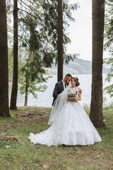Fototapeta premium A wedding couple is enjoying the best day of their lives against the backdrop of a lake and tall trees. The groom hugs the bride.