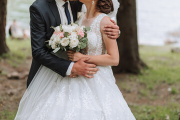 A wedding couple is enjoying the best day of their lives against the backdrop of a lake and tall trees. The groom hugs the bride.