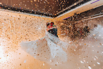 Gorgeous bride and stylish groom dancing under golden confetti at wedding reception. Happy wedding couple performing first dance in restaurant. Romantic moments