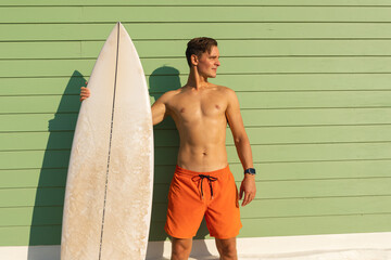 A man standing at the light green wall holding a surfboard