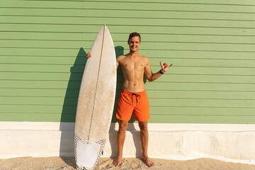 A smiling man with nice body standing at the light green wall holding a surfboard and showing shaka