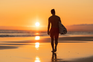 Black silhouette of an attractive man walking on the shore holding a surfboard at orange sunset