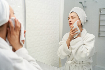 Graceful woman in a bathrobe wearing towel on head, stands in front of a mirror gently drying her face with soft towel after refreshing cleanse - 631518693