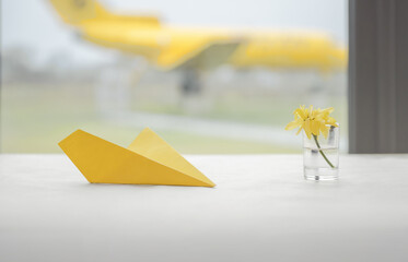 Selective focus shot of small paper airplane and yellow flower on the table in the foreground. Full...