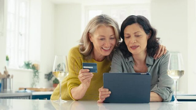 Same sex mature female couple with credit card using digital tablet at home in kitchen to make major purchase or to book holiday or tickets celebrating with glass of wine - shot in slow motion