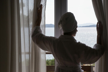Woman in a bathrobe opening curtains in room in the morning with beautiful peaceful seaview	 - obrazy, fototapety, plakaty