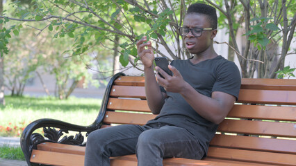 Fototapeta premium Young African Man Shocked by Loss on Smartphone while Sitting Outdoor on a Bench