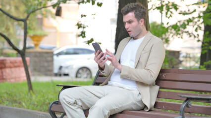 Fototapeta premium Casual Young Man Shocked by Loss on Smartphone while Sitting Outdoor on a Bench