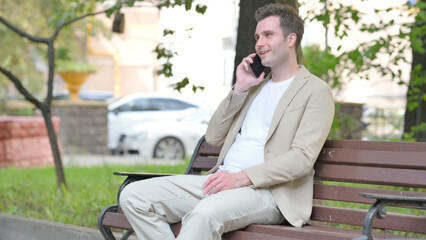 Fototapeta premium Casual Young Man Talking on phone while Sitting Outdoor on a Bench