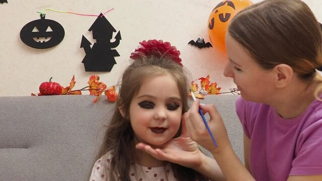 Mother making Halloween makeup to preteen girl daughter and painting spooky black lines on face. Pretty child with creepy cosmetics preparing for October holiday party