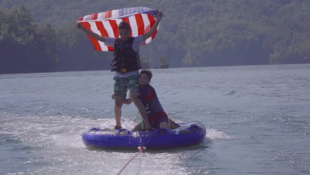 Slow-motion of two Caucasian kids with the USA flag on a donut boat floating on a lake