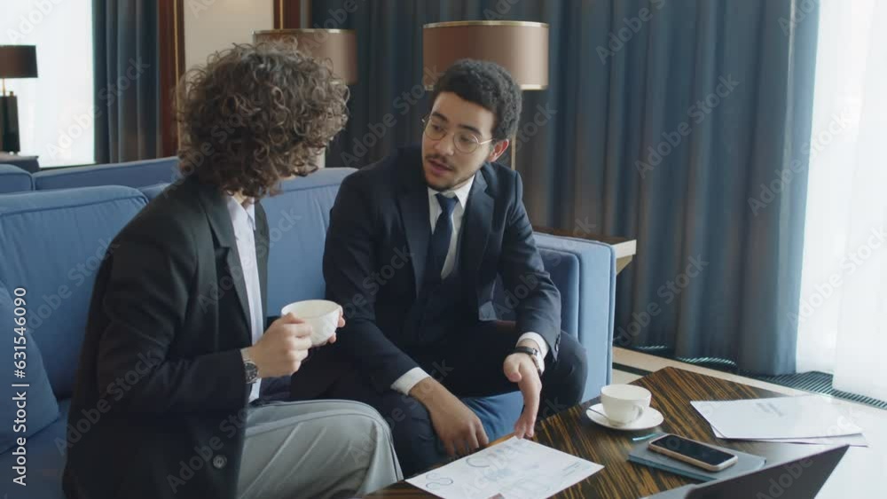 Poster medium shot of business partners discussing projects and looking at document while sitting at hotel 