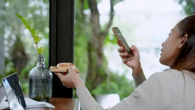 Pretty Asian woman using smartphone to take picture of bread lying on wooden tray in hand. to prepare into online store application and social media of store Instruct customers to look at it