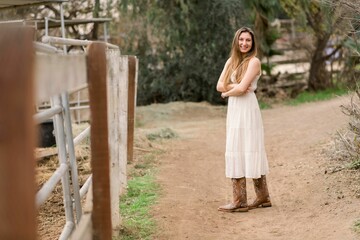 Young woman wearing a white dress and cowboy boots posing next to a wooden fence