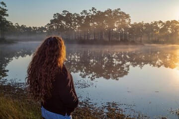 Fototapeta na wymiar Female with curly hair admiring the lake with vegetation and sunset reflection