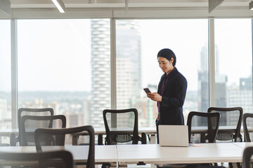Smiling Asian businessman wearing stylish black suit holding mobile phone standing in meeting room