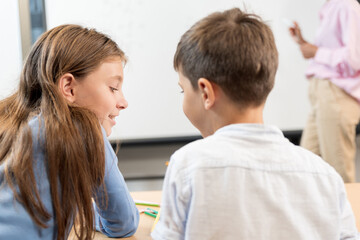 Close up, rear view portrait of schoolgirl and schoolboy. Students discuss their homework with each other. In the background, the teacher near the blackboard explains the lesson. Cheerful conversation