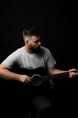 Bearded brutal guitarist plays an acoustic guitar in a black room. The concept of music recording, rehearsal or live performance.