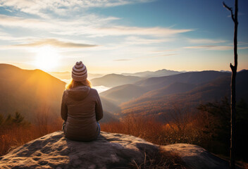Backside of a woman sitting on the mountain top in the evening or in the morning and enjoying the sunset or sunrise. - obrazy, fototapety, plakaty