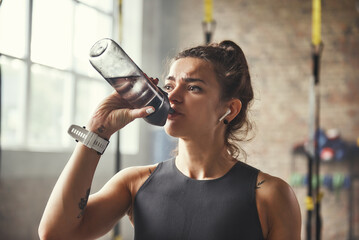 Close up of young athletic woman in earphones is drinking water while exercising in gym.