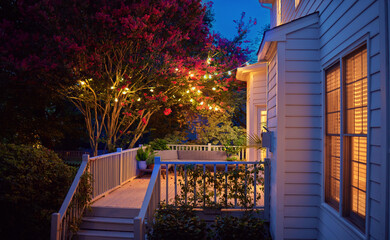beautifully lit exterior of traditional house with wooden terrace and blooming crape myrtle in summer garden at night - 631566267