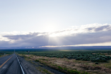 A long asphalted straight road stretches far into the horizon, on which blue mountains can be seen. The rays of the sun break through the large clouds over the road. blue sky before sun