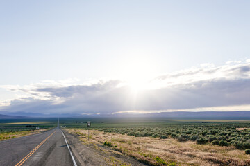 Fototapeta premium A long asphalted straight road stretches far into the horizon, on which blue mountains can be seen. The rays of the sun break through the large clouds over the road. blue sky before sun