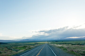 A long asphalted straight road stretches far into the horizon, on which blue mountains can be seen. The rays of the sun break through the large clouds over the road. blue sky before sun