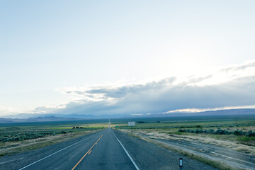 A long asphalted straight road stretches far into the horizon, on which blue mountains can be seen. The rays of the sun break through the large clouds over the road. blue sky before sun