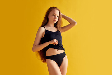 Portrait. Horizontal image of redhead woman with slim body, looking at camera in black underwear against bright yellow background.