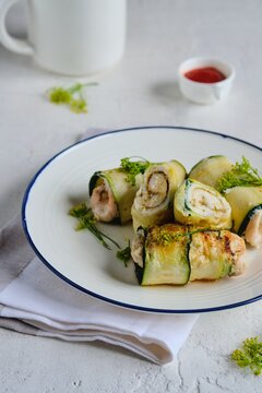 Fried chicken rolls wrapped with sliced zucchini on a white plate on a light concrete background. Recipes for zucchini.
