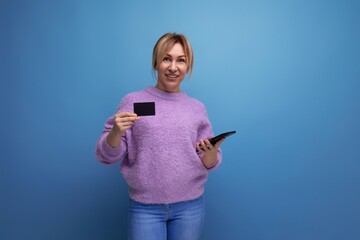 cute blonde young woman in purple hoodie holding credit plastic card mockup and smartphone on blue background with copy space