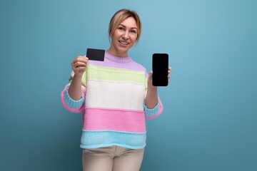 attractive blond woman in casual look demonstrating mockup of credit card and smartphone on blue...
