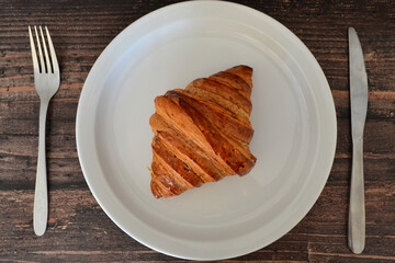 Croissant on white plate with fork and knife on wooden table