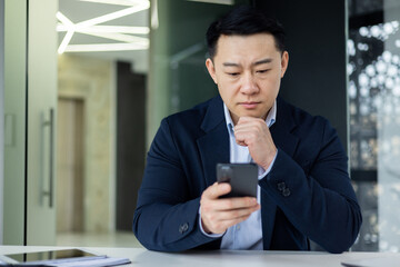 Mature thinking asian boss inside office workplace with phone in hands, successful businessman thinking sitting with laptop in business clothes.